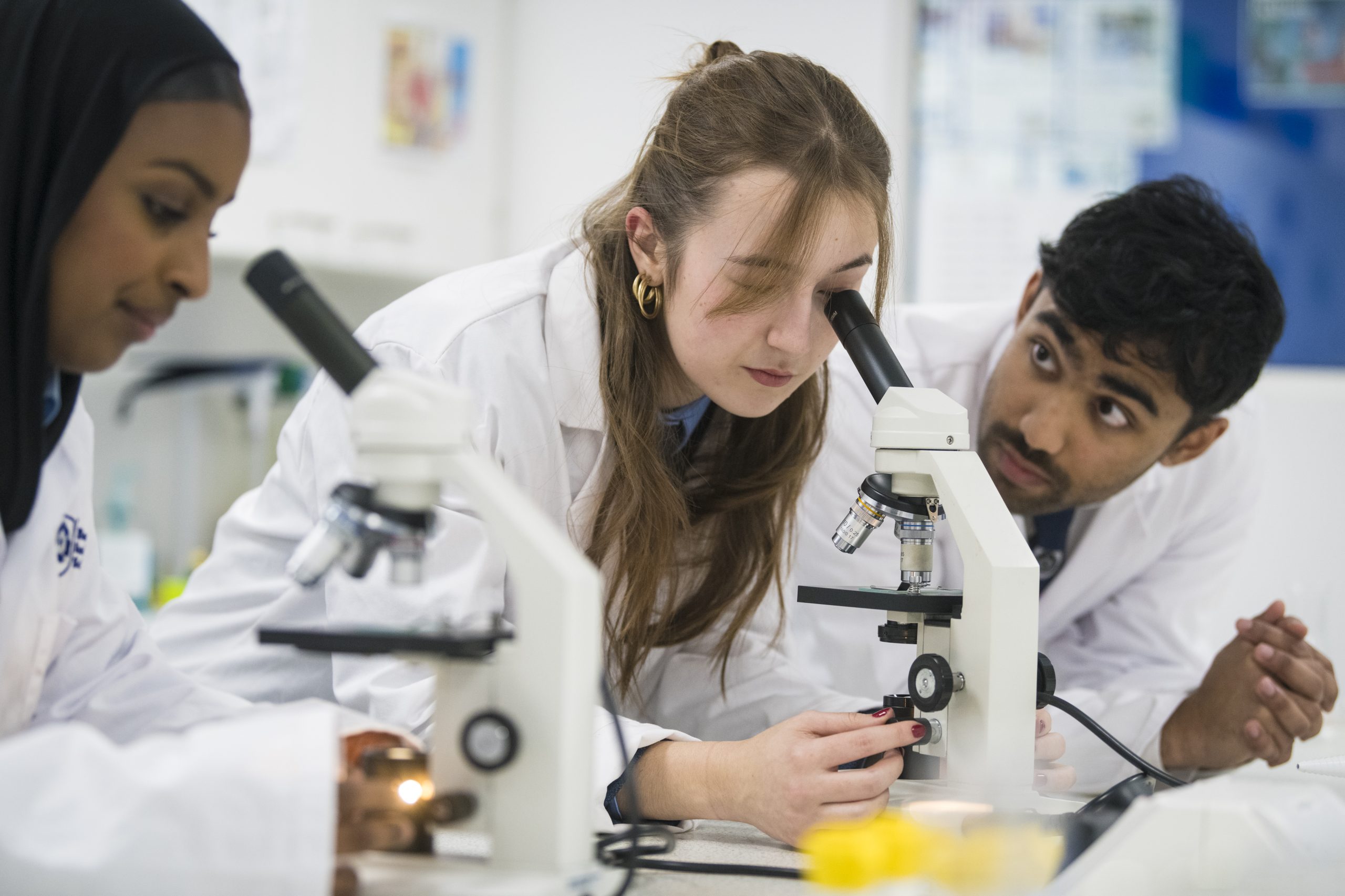 Two female students and one male student in lab coats using microscopes